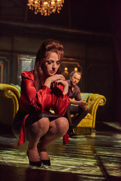 Scene from Bat Out Of Hell The Musical : Sloane (Sharon Sexton) crouches pensively in the foreground, behind her Falco (Rob Fowler) sits on a gaudy looking couch, looking in her direction