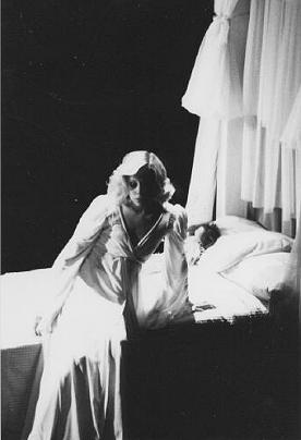 Ellen Foley as Wendy, dressed in all white and looking contemplative as she sits on the edge of her bed.